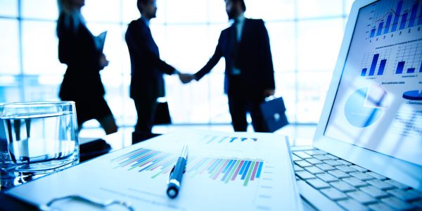 Laptop, financial document with pen and glass of water at workplace on background of three business partners making a deal